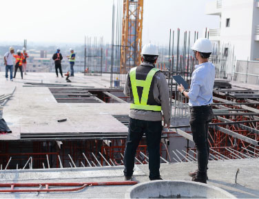 Two construction workers in hard hats stand on a construction site, surrounded by building materials and machinery.